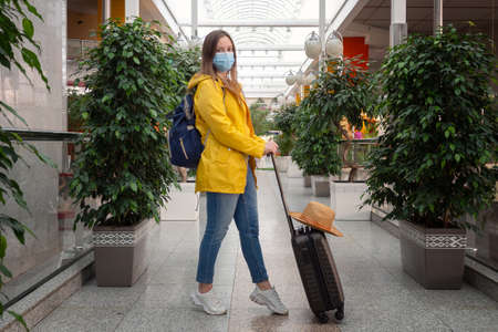 happy modern tourist woman in yellow with face medical mask and luggage at the airport station. happiness to travel again, opening borders for tourism. new normal for travelersの写真素材