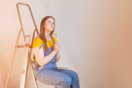 A pensive woman with brush and ladder looks dreamily in a room with an unpainted wall with copy space. Independent single female makes DIY repairs in her apartment.の写真素材