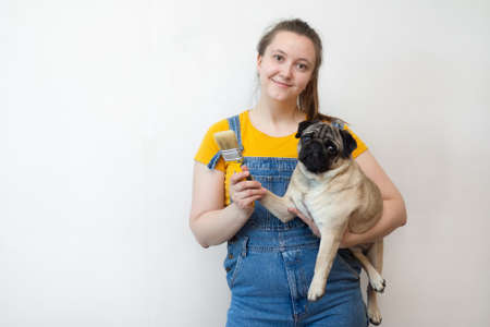 woman holding pet pug dog with paint brush showing wall with copy space. Independent single female makes DIY repairs in her apartment. place for textの写真素材