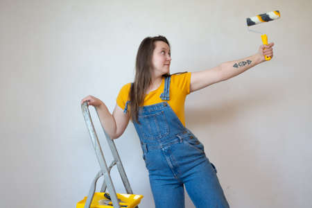 Young happy woman makes repairs in her apartment. Funny female with a roller on ladder. Independent single female with tattooの写真素材