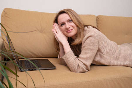 Woman lying on a sofa at home and working on a laptop computer. Cozy remote work. online consultation and psychotherapy session. looking at the cameraの写真素材