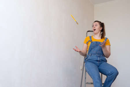 Happy woman playing with brush on ladder in a room with an unpainted wall with copy space. Independent single female makes DIY repairs in her apartment.の写真素材