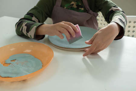 child painting with sponge on clay pottery plate in ceramic workshop studio. Development of art and painting in children. Close upの写真素材