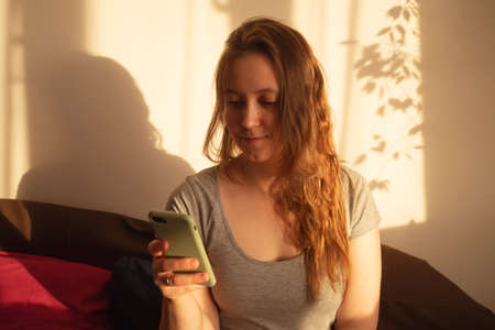 Young woman looking at smartphone in warm rays of sunset. Warm calm portrait of girl indoor. light and shadow on the face.の写真素材