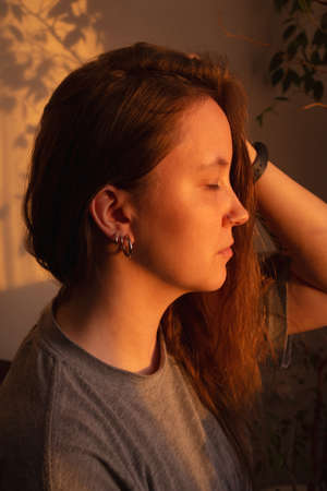Young woman close eyes and relaxation in warm rays of sunset. Warm calm portrait of girl indoor