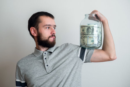 man holding big glass jar with money banknotes on hand. finance power and stability concept. moderate consumption and economyの写真素材