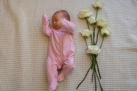 Caucasian infant girl in pink bodysuit lying on her back with roses flowers on the bed at home....