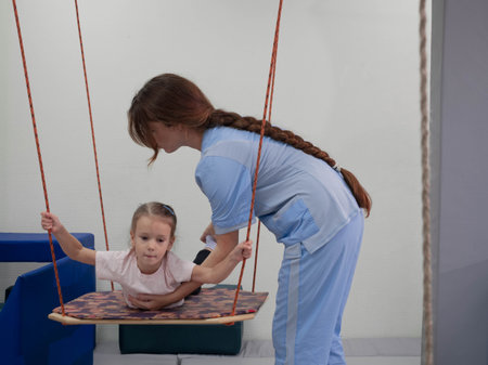 Child with physiotherapist on swing during sensory integration session. Little girl in rehabilitation centre doing exercises for development functions of the vestibular apparatusの写真素材