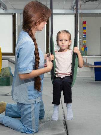 Child with physiotherapist on swing during sensory integration session. Little girl in rehabilitation centre doing exercises for development functions of the vestibular apparatusの写真素材