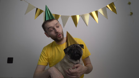 Caucasian man owner congratulation cute funny pug dog with pet birthday in a party hat. Domestic animal love and pampering concept. Celebrating pets birthday enjoysの写真素材