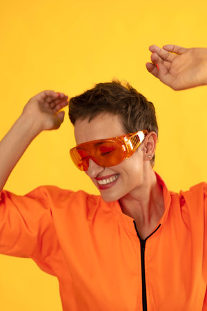 Middle age funny and Cheerful woman wear modern Orange overalls with glasses standing over yellow color background. Female having fun and energetic movement studio shotの写真素材