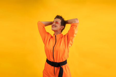 Cheerful woman with colorful make up wear modern Orange overalls posing over yellow color background. Middle age happy female having fun and energetic movement with copy spaceの写真素材