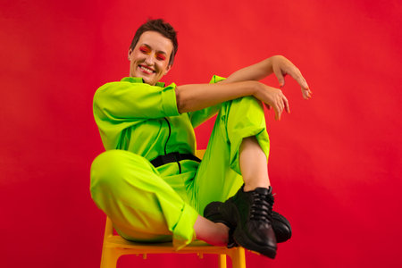 Middle age funny and Cheerful woman wear modern green overalls sitting on chair over red color background. Full length body size photo Female having fun and energetic movement studio shotの写真素材