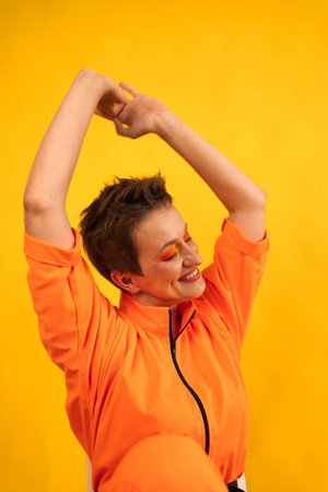 Middle age cheerful woman with colored make up wear modern Orange overalls and sitting on white chair over yellow color background. Female having fun and energetic movement studio shotの写真素材