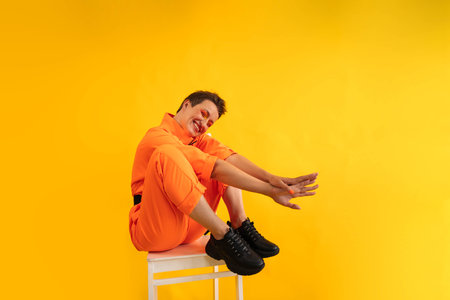 Middle age funny and Cheerful woman wear modern Orange overalls sitting on white chair over yellow color background. Female having fun and energetic movement studio shotの写真素材