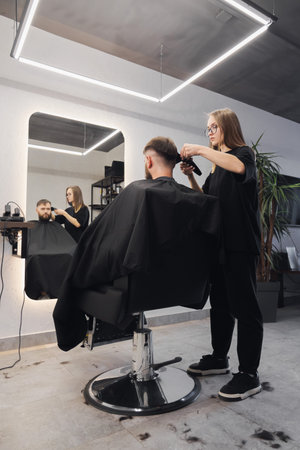 Professional woman barber wielding electric hair clipper making fashionable haircut. Bearded man client visit gentleman grooming beauty salon for hairstyle care. Modern haircut style in barber shopの写真素材