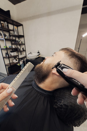 Barber wielding electric hair clipper shaving beard customer in modern barbershop. Bearded Man client visit gentleman grooming beauty salon for hairstyle care. Male beard style conceptの写真素材