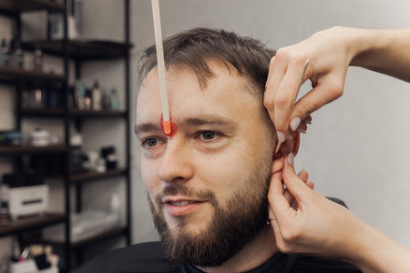 Barber removing hair in ears and eyebrow by waxing from Bearded Man face in stylish barbershop. Beautician hair removal procedure in male beauty salon. Male skin care and beard style conceptの写真素材