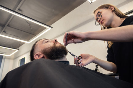 Woman Barber shaving bearded customer in modern style barbershop. Male Beard hair cutting with scissors. Man client visit gentleman grooming beauty salon for evens the beardの写真素材