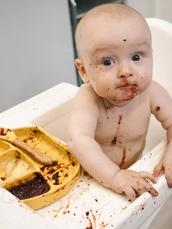 Toddler covered in food sits in a high chair and enthusiastically eating for meal. Concept of early childhood nutrition and introducing solid foods to infants.の写真素材