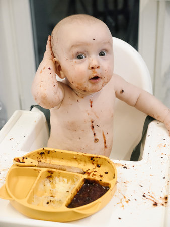 Toddler covered in food sits in a high chair and enthusiastically eating for meal. Concept of early childhood nutrition and introducing solid foods to infants.の写真素材