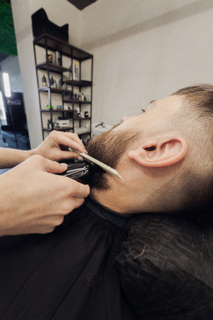 Woman Barber wielding electric hair clipper shaving beard customer in modern barbershop. Bearded Man client visit gentleman grooming beauty salon for hairstyle care. Male beard style conceptの写真素材