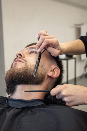 Barber shaving bearded customer in modern style barbershop. Male Beard hair cutting with scissors close up. Man client visit gentleman grooming beauty salon for evens the beardの写真素材