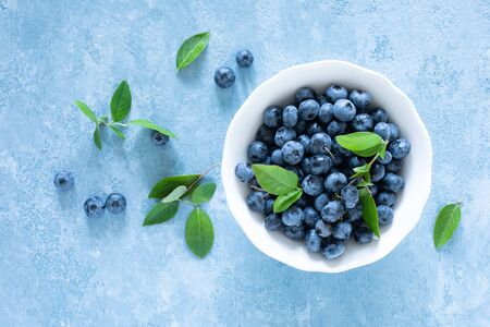 Blueberries with leaves in white bowl on blue background, top view