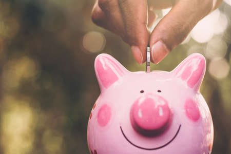 A coin in the hand of a man who is putting it in a pink piggy bank.の写真素材