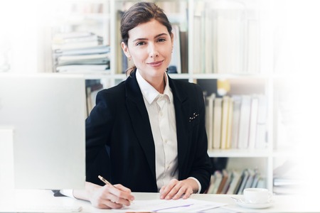 Portrait of smiling businesswoman looking at camera and smiling while working with documents at office in front of computerの写真素材