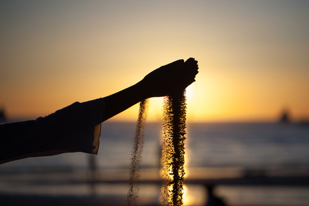 Silhouette of a young girl's hand pouring sand from a beach against a yellow sky at sunsetの写真素材