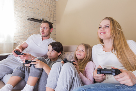 Family of parents and two children playing video game together sitting on sofa at home