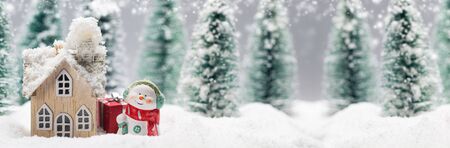 Small decorative snowman with gift near wooden house in fir forest under falling snowの写真素材
