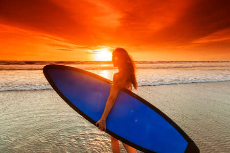 Beautiful mixed race woman on tropical beach holding surfboard at sunsetの写真素材