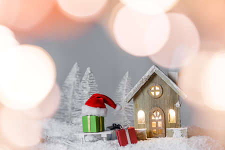 Christmas card with small glowing decorative house, gifts and Santa Claus cap on sleigh on snow over fairy tale winter fir forest background and falling snow. New Year design postcard. Copy space for textの写真素材