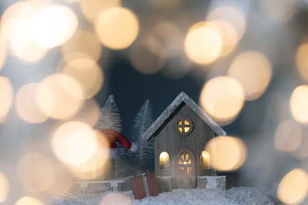 Christmas card with small glowing decorative house, gifts and Santa Claus cap on sleigh on snow over fairy tale winter fir forest background and falling snow. New Year design postcard. Copy space for textの写真素材
