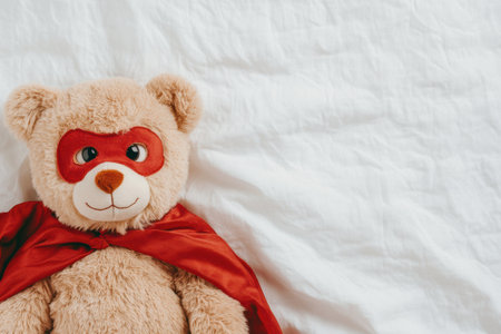 Cute plush Bear in Red Superhero Costume Lying on Bed with White Sheets.の素材