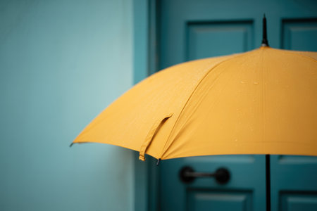 Bright yellow umbrella covered with raindrops leaning against a turquoise door on a rainy day with soft ambient light.の素材