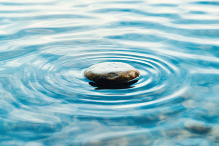 Smooth stone creating gentle ripples in clear blue water during calm daylight, focusing on tranquility and natural balance.の素材