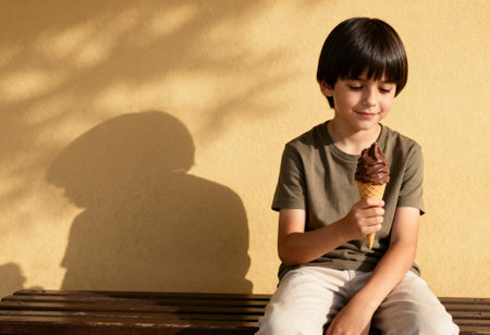 Smiling boy enjoys chocolate ice cream cone sitting on wooden bench against yellow wall in warm afternoon sunlight.の素材
