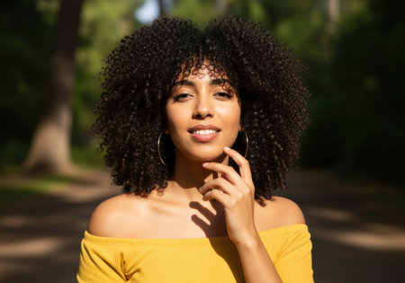 Confident young woman with natural curly hair in sunlight wearing yellow off shoulder top standing outdoors in green park.の素材