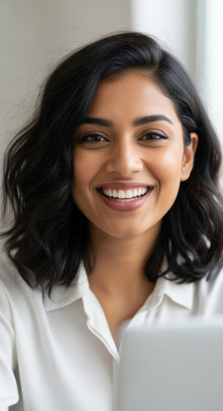 Confident young woman with wavy black hair smiling brightly while working on a laptop in a bright indoor office space.の素材