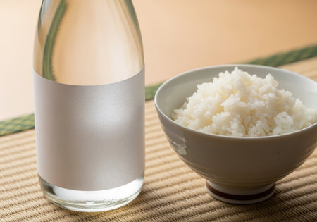 Clear glass bottle with blank label beside a ceramic bowl of steamed white rice on a traditional woven mat in soft lighting.の素材