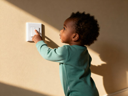 Curious toddler in green pajamas reaching to turn off a light switch on a beige wall in a sunlit modern home interior.の素材