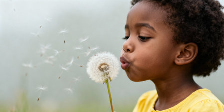Young child blowing dandelion seeds outdoors, creating a whimsical moment in nature with soft background and warm lighting.の素材