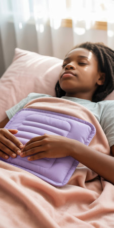African american girl resting in bed with eyes closed, holding a purple heating pad on her stomach under soft natural light.の素材