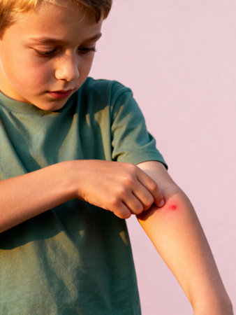 Young boy inspecting insect bite on arm against soft pink background with curious expression and visible skin irritation.の素材
