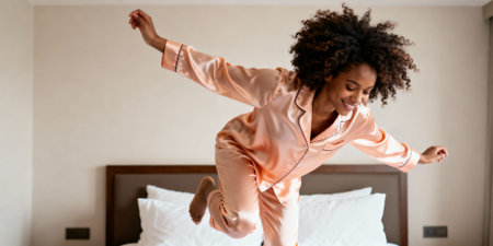 Joyful young woman wearing satin pajamas jumping on a bed in a modern bedroom filled with soft morning sunlight and positive energy.の素材