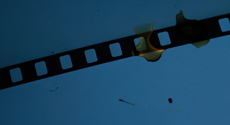Damaged film strip with burn marks isolated on a blue background under moody lighting, symbolizing loss and deterioration.の素材