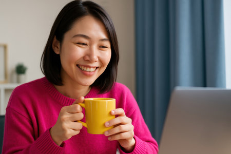 Smiling young woman in pink sweater enjoying coffee while working on laptop indoors during the morning at home.の素材